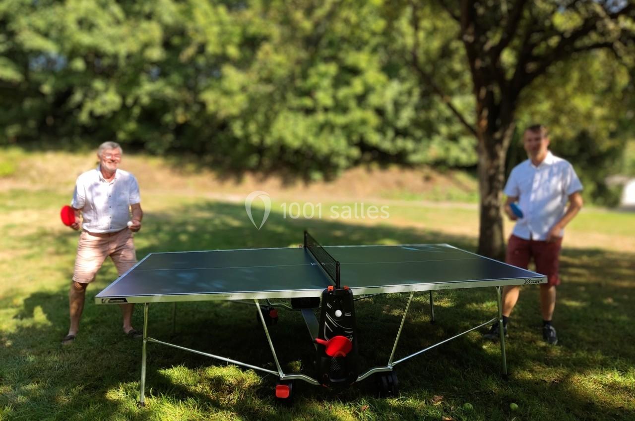 Deux hommes jouent au tennis de table en plein air dans un jardin ombragé par des arbres.