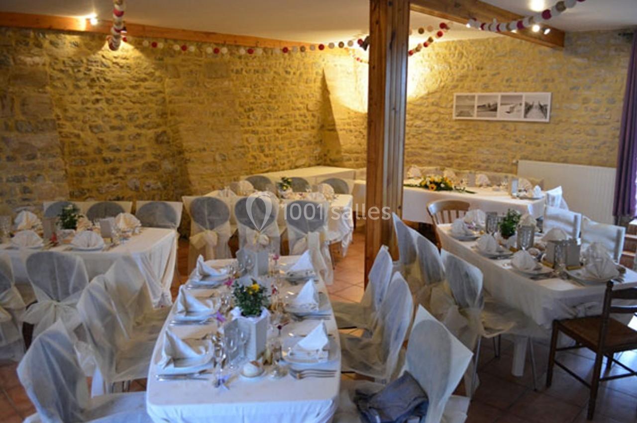 Salle décorée pour un événement avec des tables dressées, nappes blanches, chaises couvertes et murs en pierre apparente.