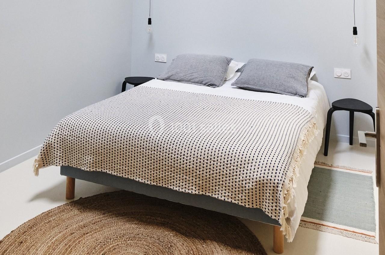 Lit double avec couverture à motifs, deux oreillers gris, tables de chevet noires et tapis rond en jute dans une chambre…