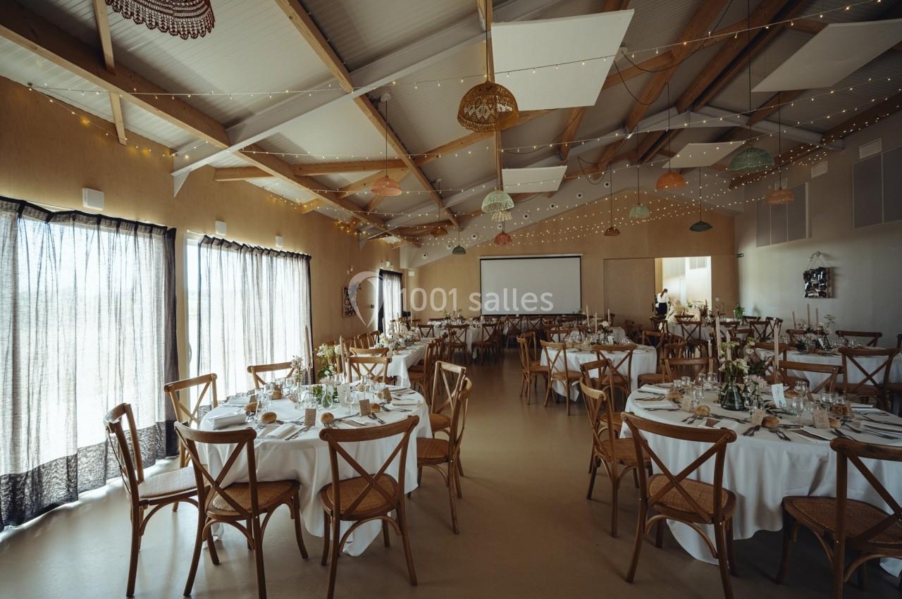 Salle de réception décorée avec des tables rondes dressées, des chaises en bois et des guirlandes lumineuses suspendues.