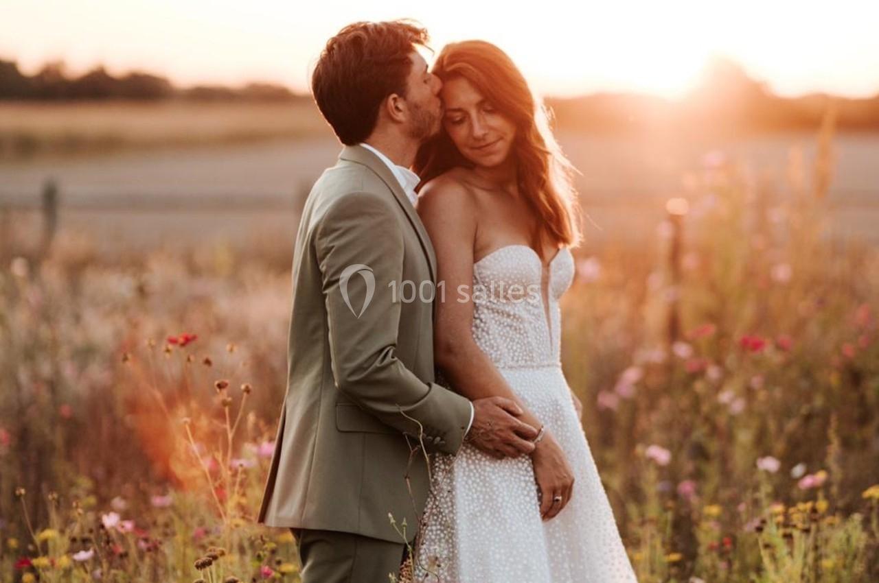 Un couple en tenue de mariage pose dans un champ fleuri au coucher du soleil.