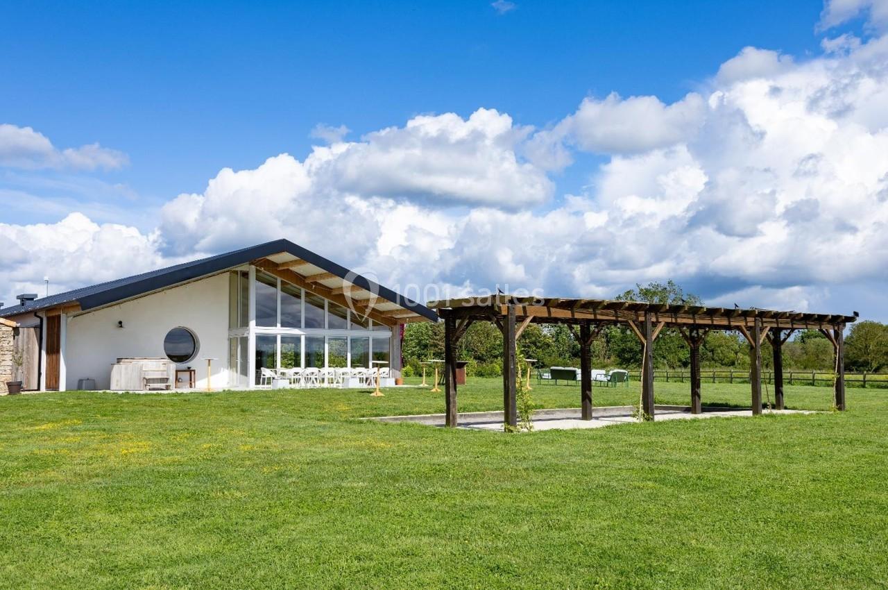 Maison moderne avec façade vitrée, pergola en bois et grand jardin verdoyant sous un ciel partiellement nuageux.