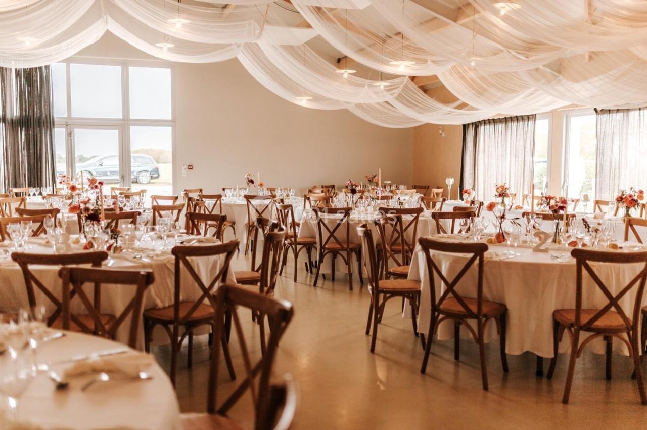 Salle de réception décorée avec des tables rondes, nappes blanches, chaises en bois et voilages suspendus au plafond.