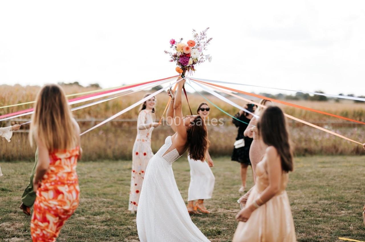 Une femme en robe blanche lance un bouquet de fleurs, entourée de personnes dans un champ avec des rubans colorés.