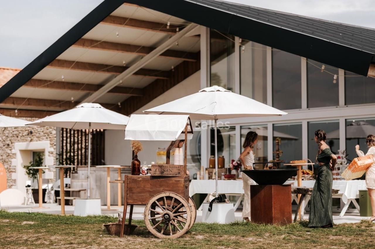 Un stand en bois avec parasols blancs devant un bâtiment moderne, entouré de personnes lors d'un événement en plein air.
