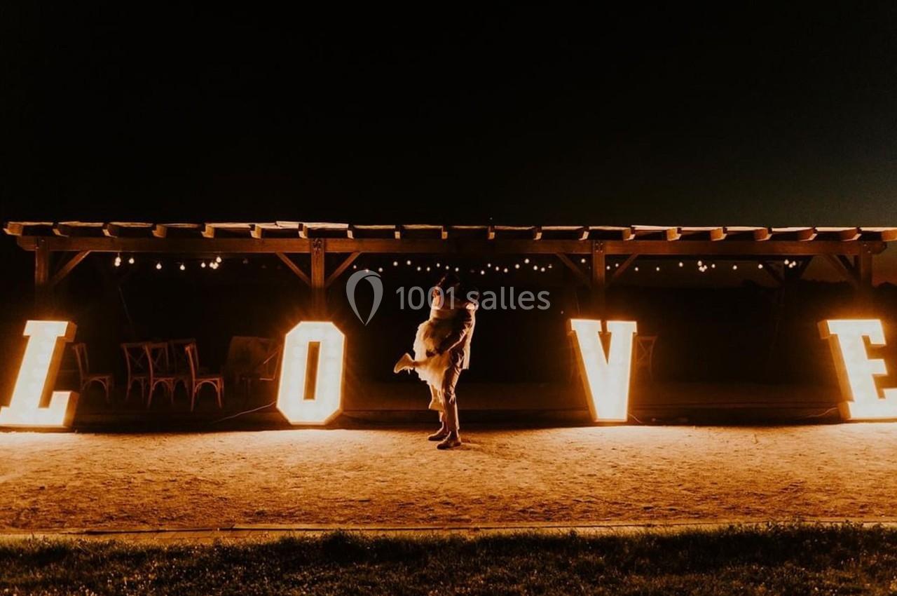 Un couple s'enlace devant de grandes lettres lumineuses formant le mot ’LOVE’ dans un décor nocturne.