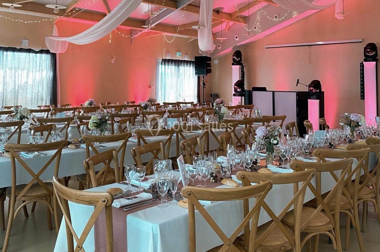 Salle décorée pour un événement, avec tables dressées, chaises en bois, nappes blanches et éclairage rose.