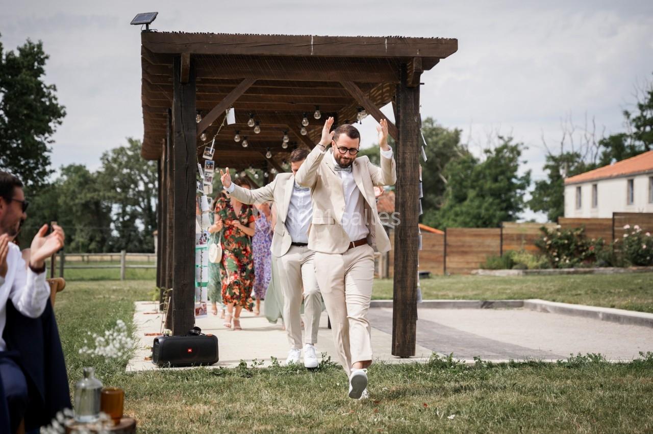 Un homme en costume beige danse joyeusement sous une pergola décorée, suivi par d'autres personnes lors d'une fête en…