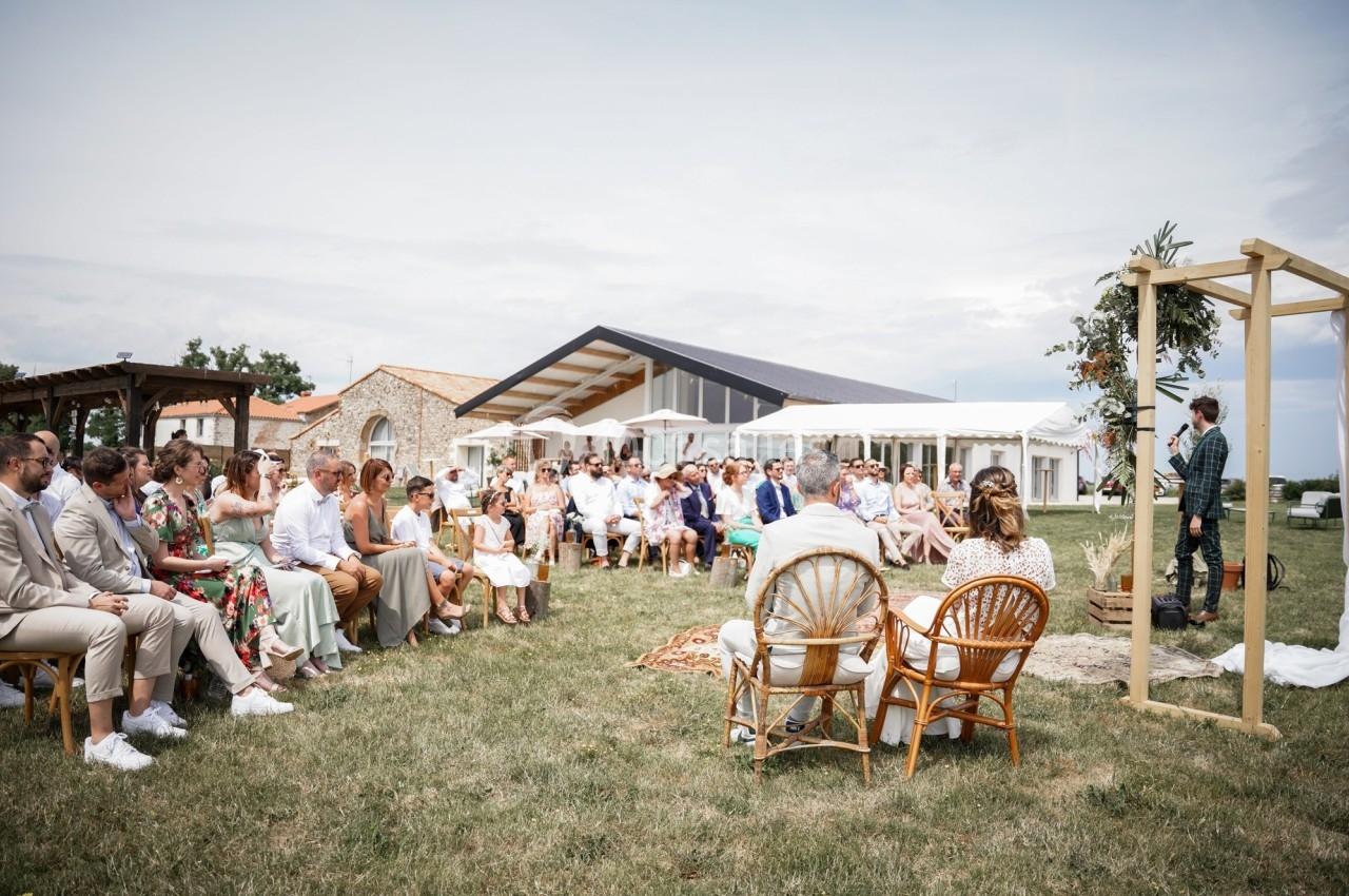 Un groupe de personnes assises en extérieur lors d'une cérémonie, avec une arche décorée et un bâtiment en arrière-plan.
