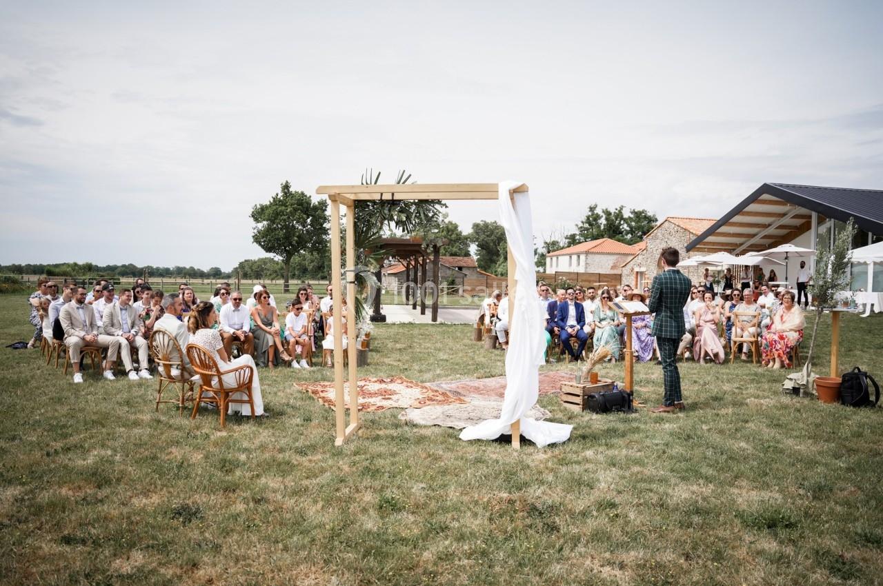 Cérémonie en plein air avec des invités assis, un officiant et une arche décorée sur une pelouse.