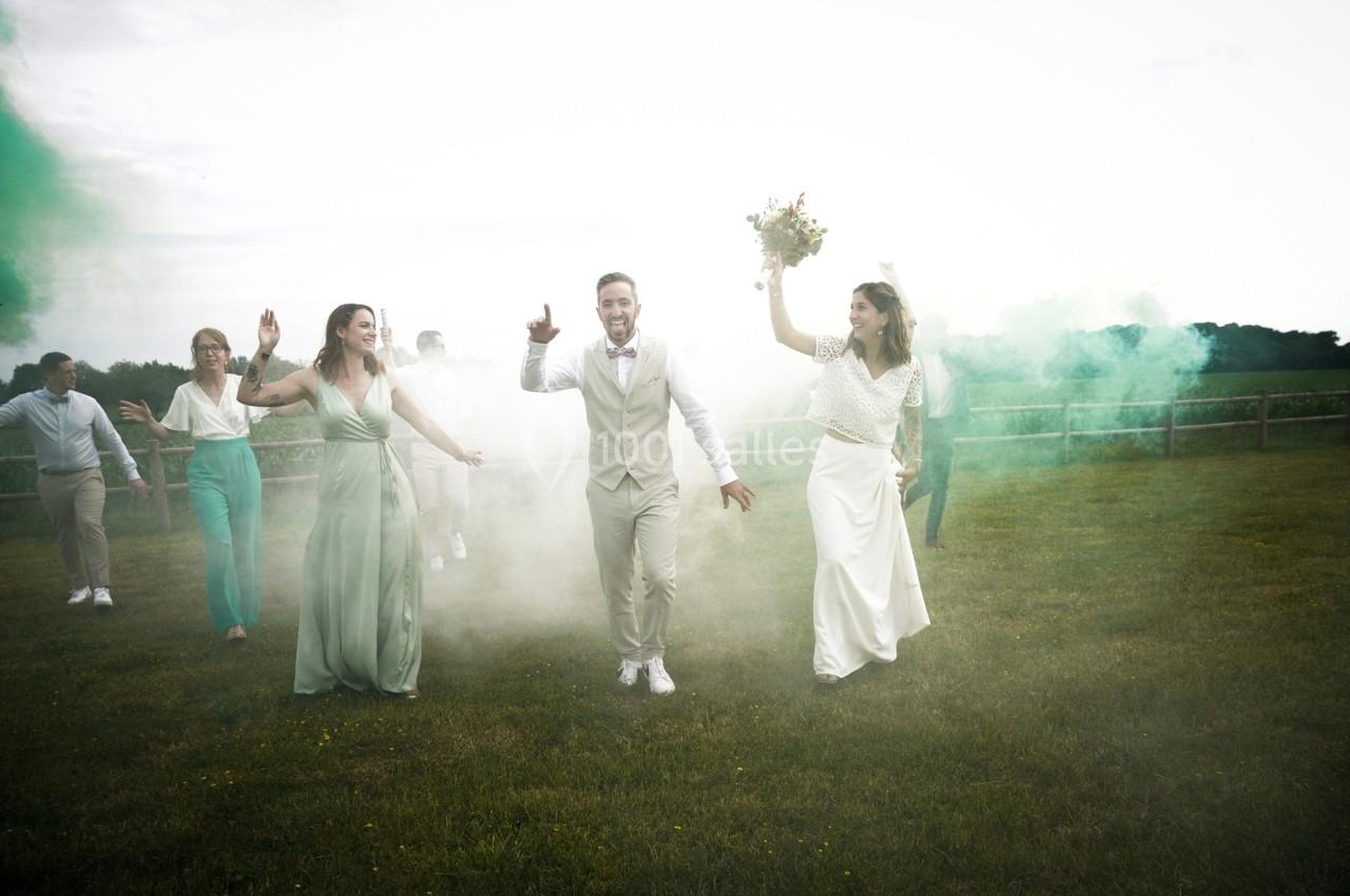 Un couple de mariés danse joyeusement avec leurs invités dans un champ entouré de fumée colorée verte.