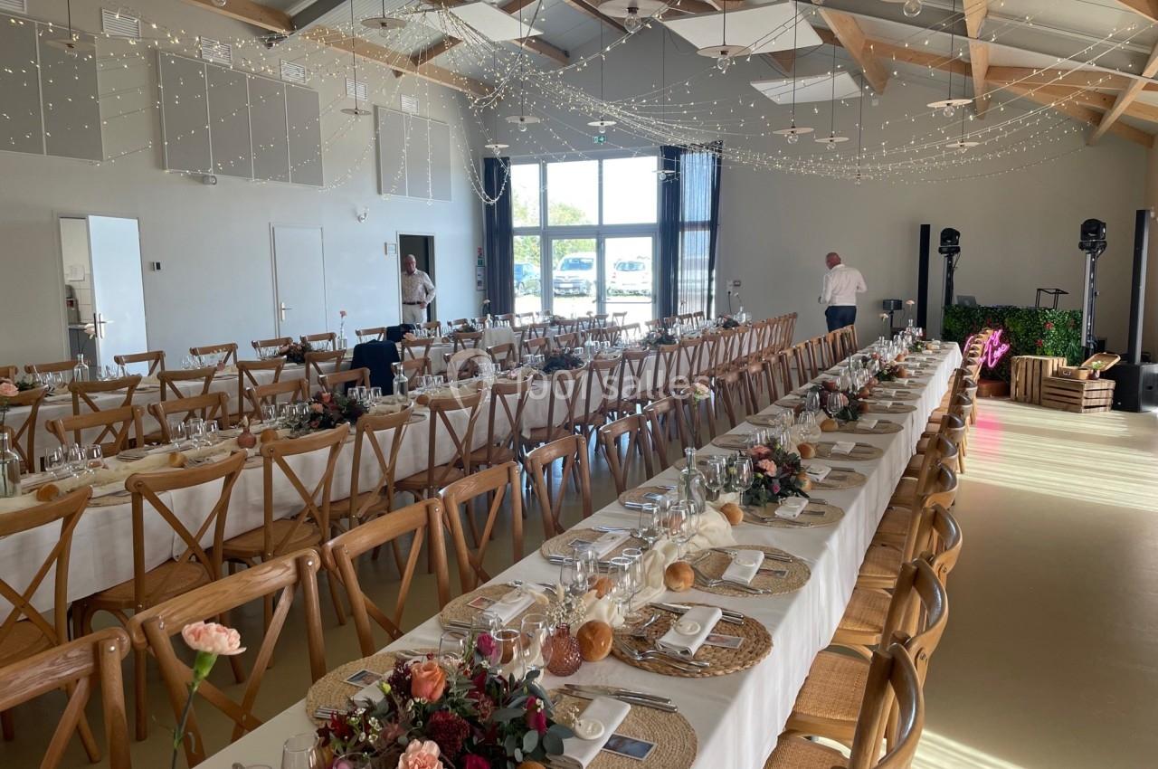 Salle de réception décorée pour un événement, avec longues tables dressées, chaises en bois et guirlandes lumineuses…