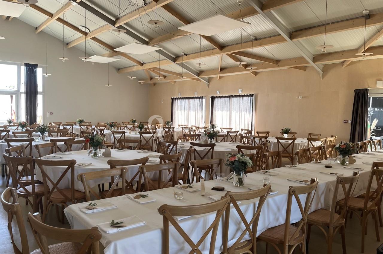Salle de réception lumineuse avec des tables dressées, nappes blanches, chaises en bois et décorations florales.