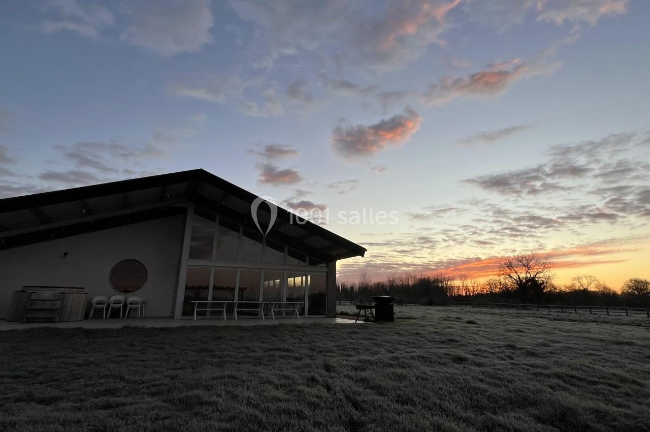 Maison moderne avec grande baie vitrée, entourée d'herbe givrée, sous un ciel de lever de soleil nuageux.