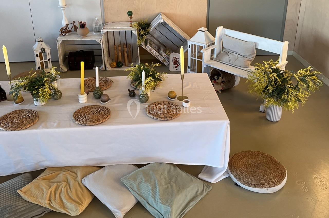 Table décorée avec des bougies, fleurs et sets en osier, entourée de coussins, bancs et éléments rustiques en bois.