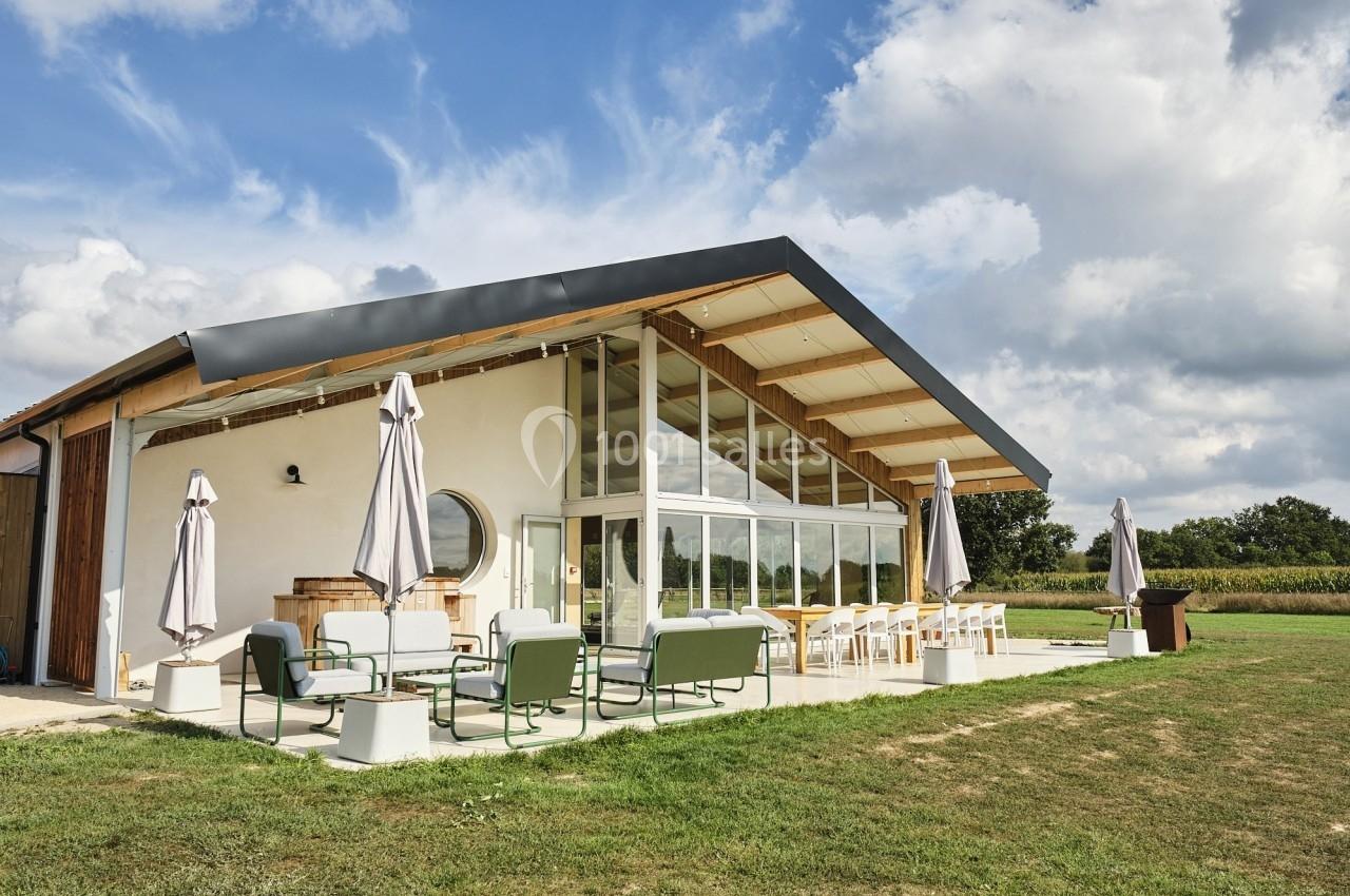 Maison moderne avec façade vitrée, terrasse aménagée avec mobilier et parasols, entourée de pelouse et ciel dégagé.