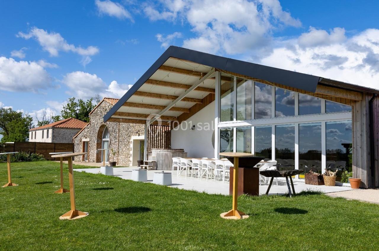 Salle moderne avec façade vitrée, terrasse aménagée et tables hautes, entourée de pelouse et ciel dégagé.