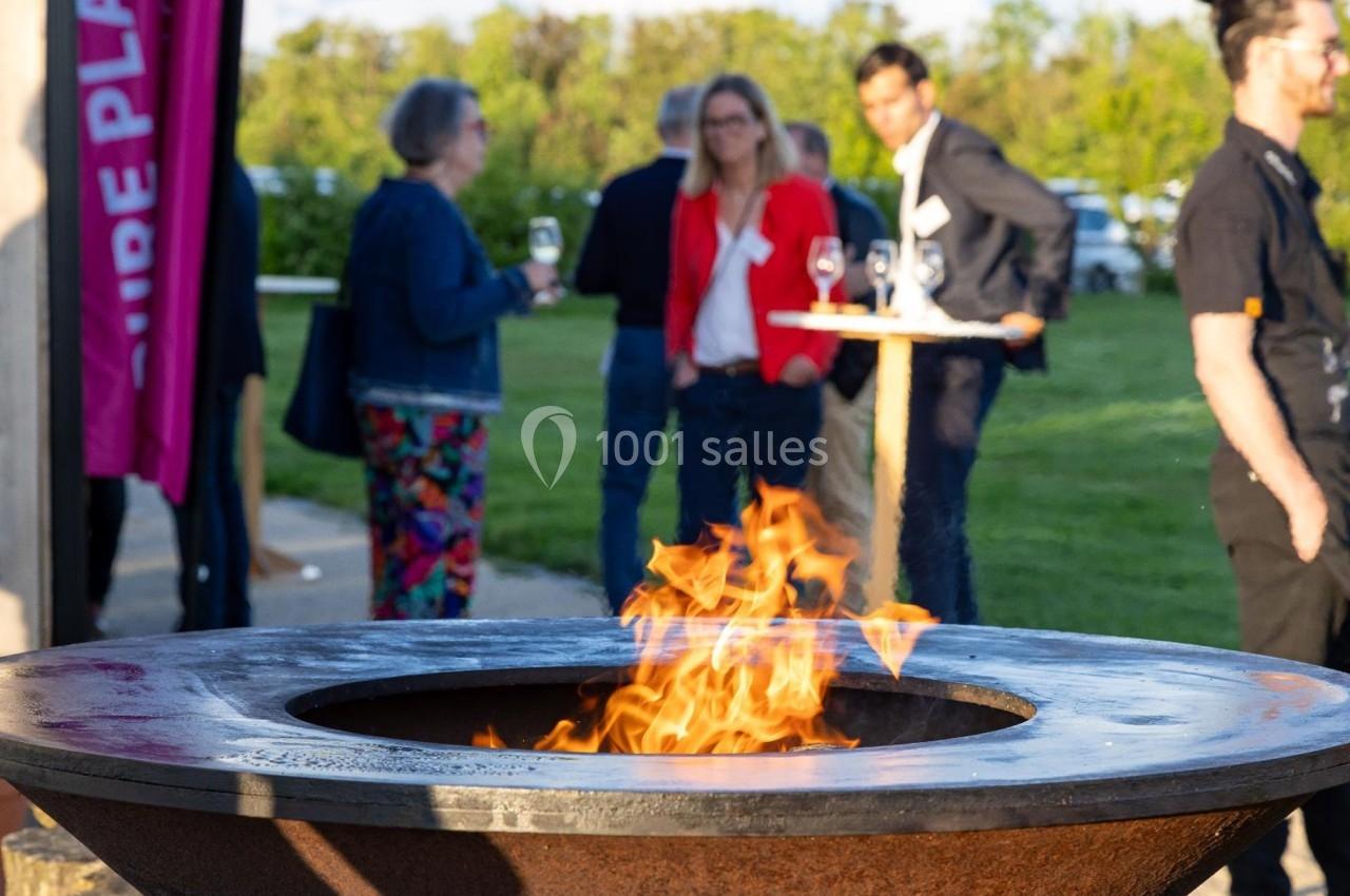 Brasero allumé au premier plan, avec des personnes discutant et tenant des verres en arrière-plan dans un espace extérieur.
