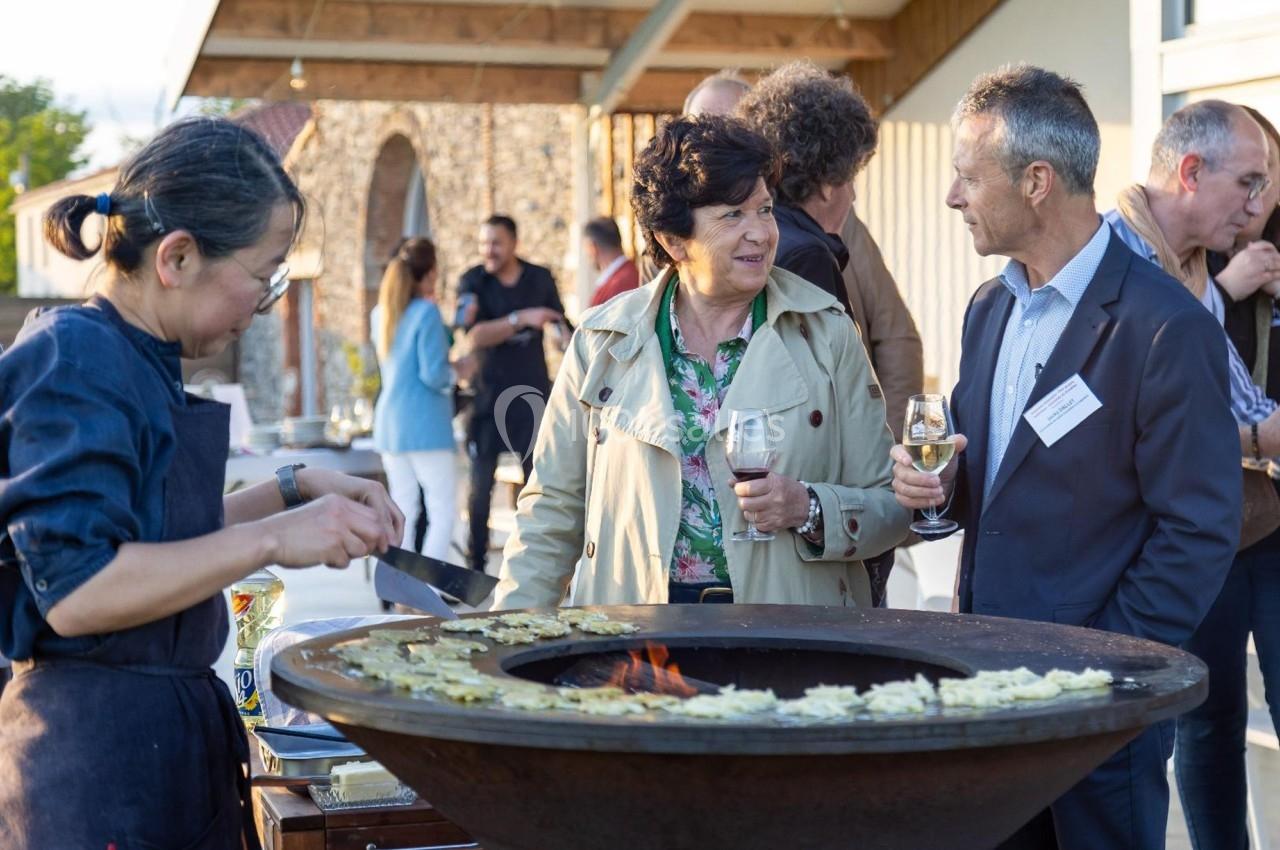 Un chef cuisine des plats sur une plancha ronde lors d'un événement en plein air avec des convives discutant.