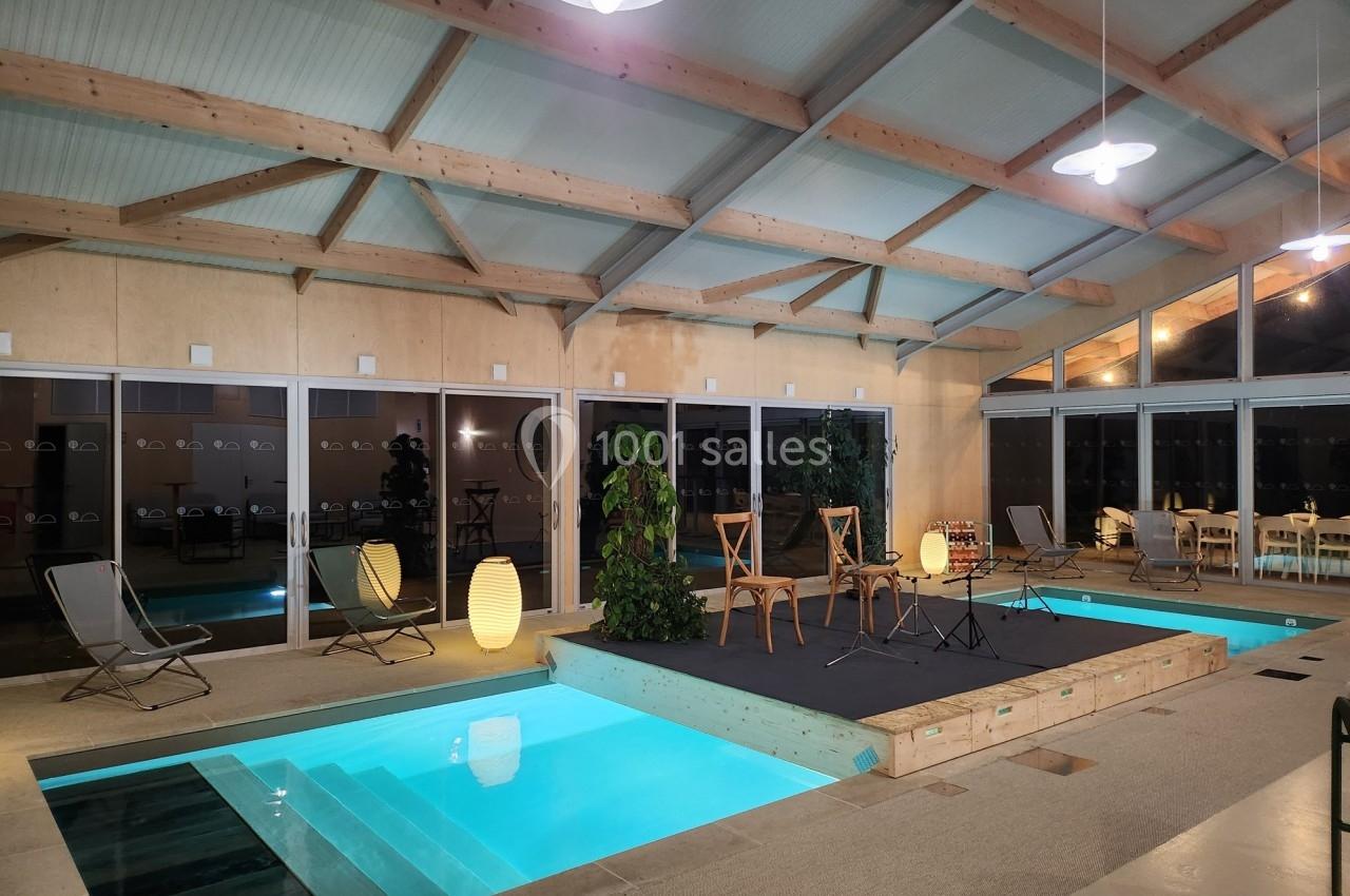 Piscine intérieure avec éclairage tamisé, entourée de chaises, plantes et d'une estrade en bois sous une structure moderne.