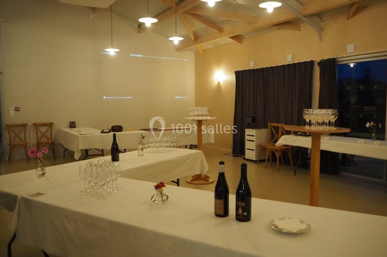 Salle lumineuse avec tables dressées, verres à vin, bouteilles et décoration simple, prête pour une réception.