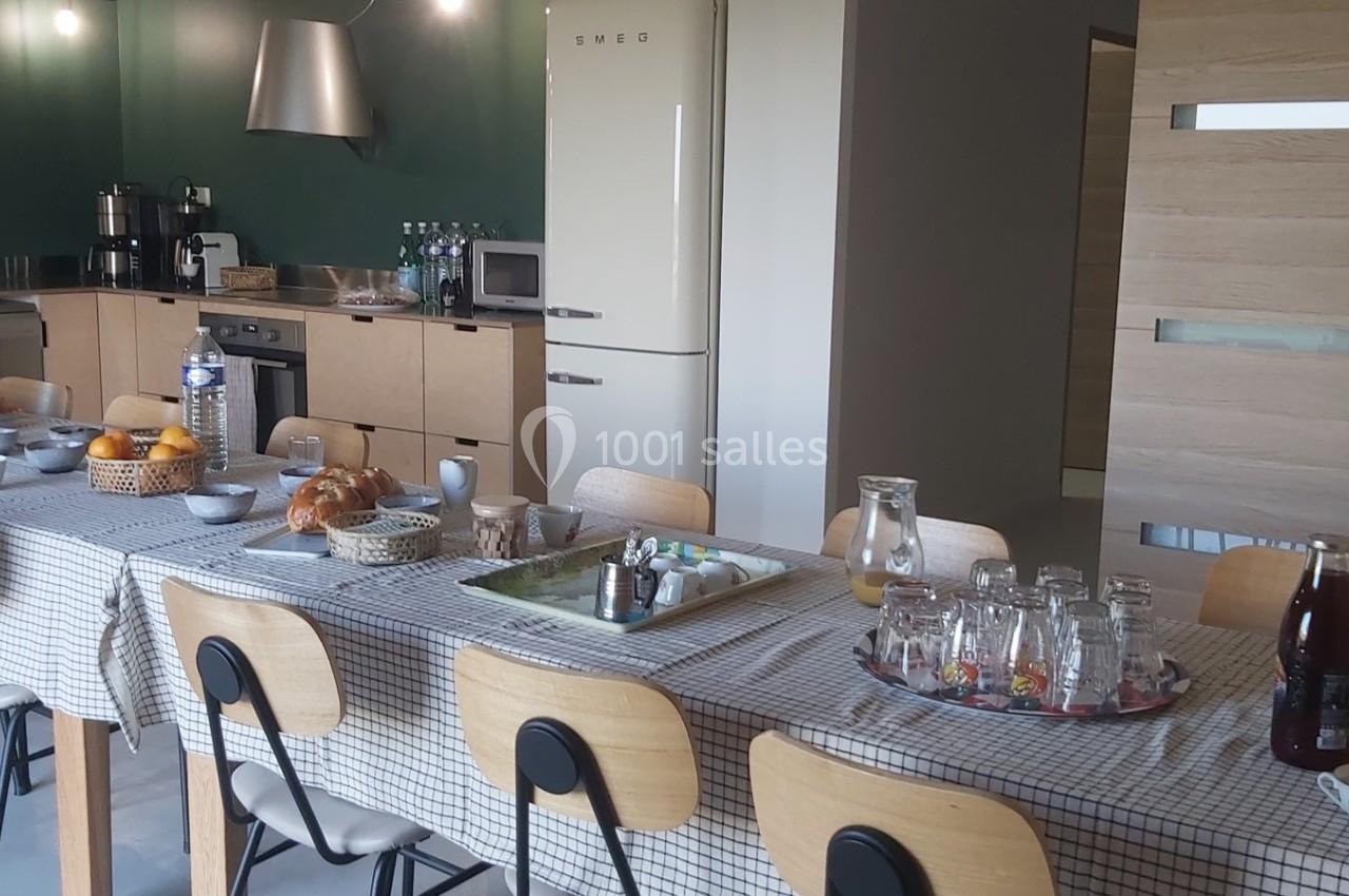 Grande table dressée avec nappes à carreaux, petits-déjeuners servis, dans une cuisine moderne avec équipements visibles.