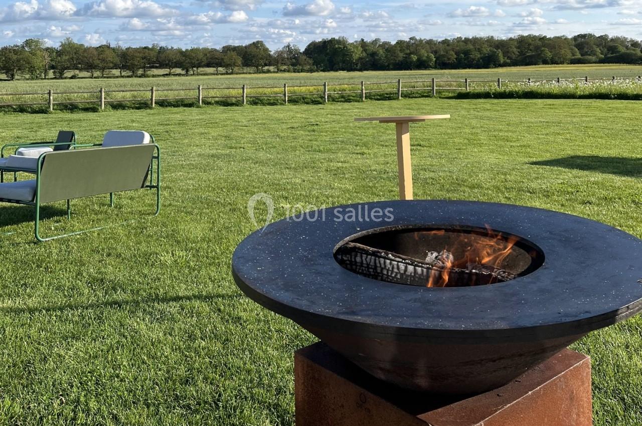 Brasero allumé au premier plan sur une pelouse, avec des sièges et une table en arrière-plan dans un cadre champêtre.