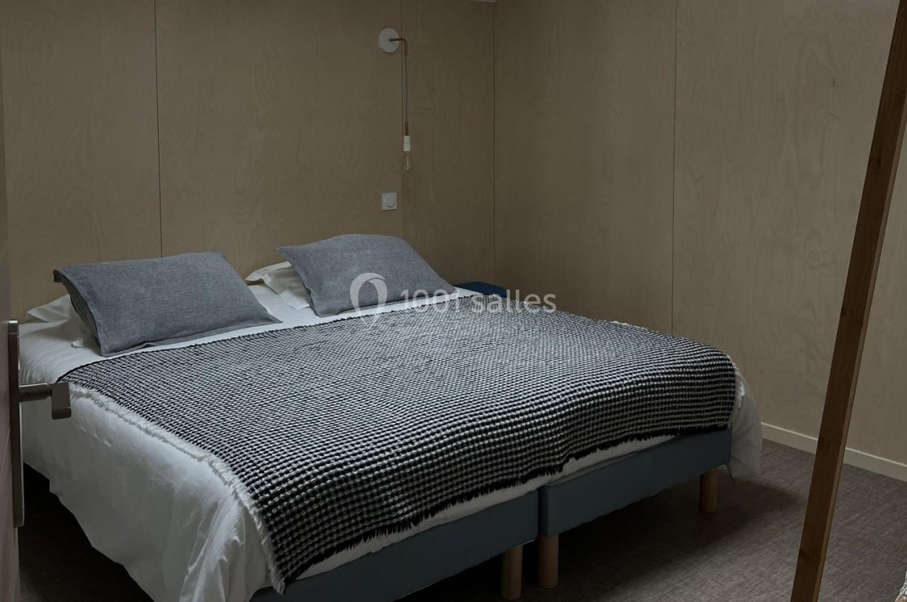 Lit double avec draps blancs, plaid à motifs noirs et blancs, et coussins gris dans une chambre aux murs en bois clair.