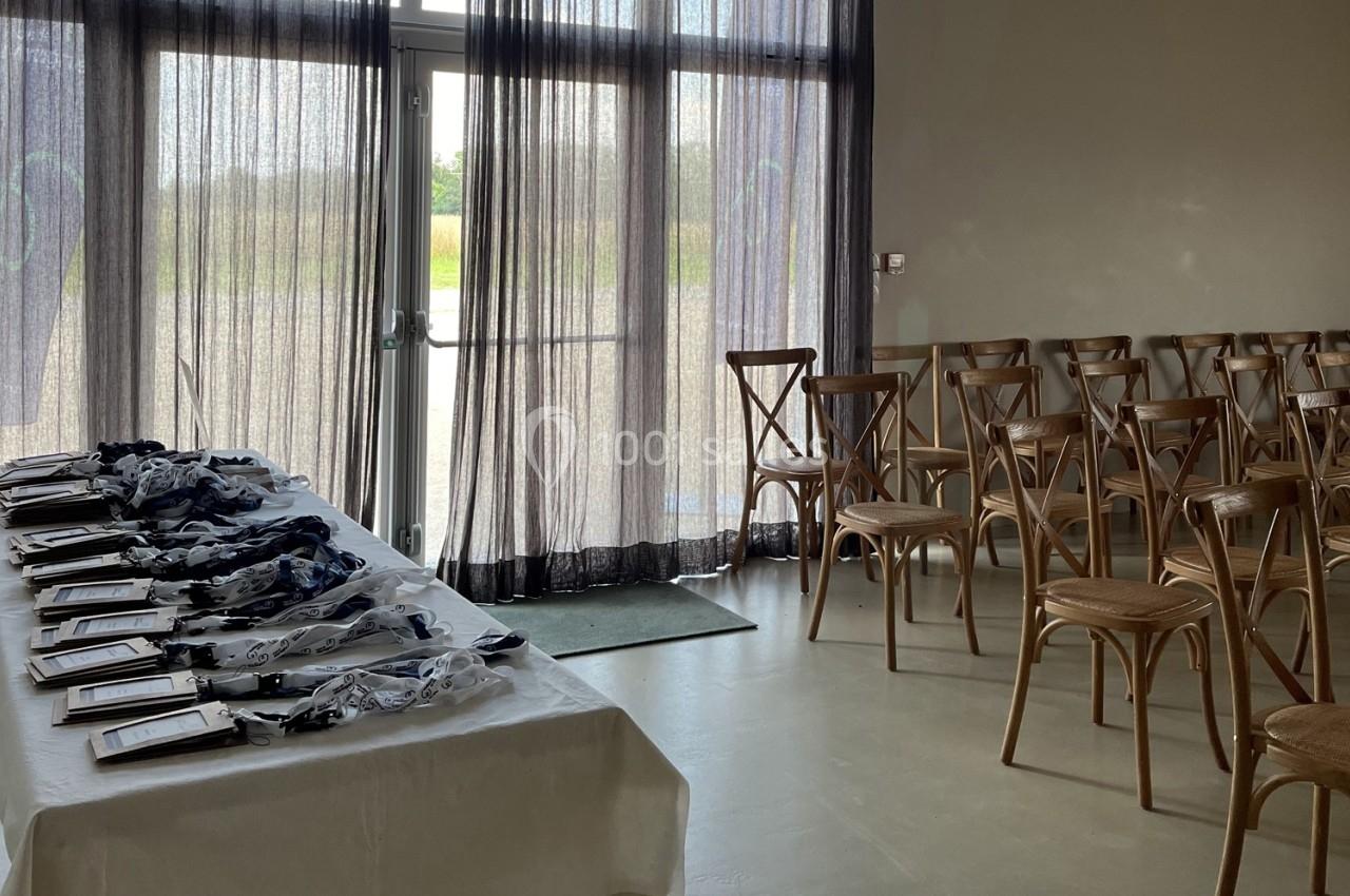 Salle avec des chaises en bois alignées et une table dressée contenant des médailles et des cadres près d'une baie vitrée.