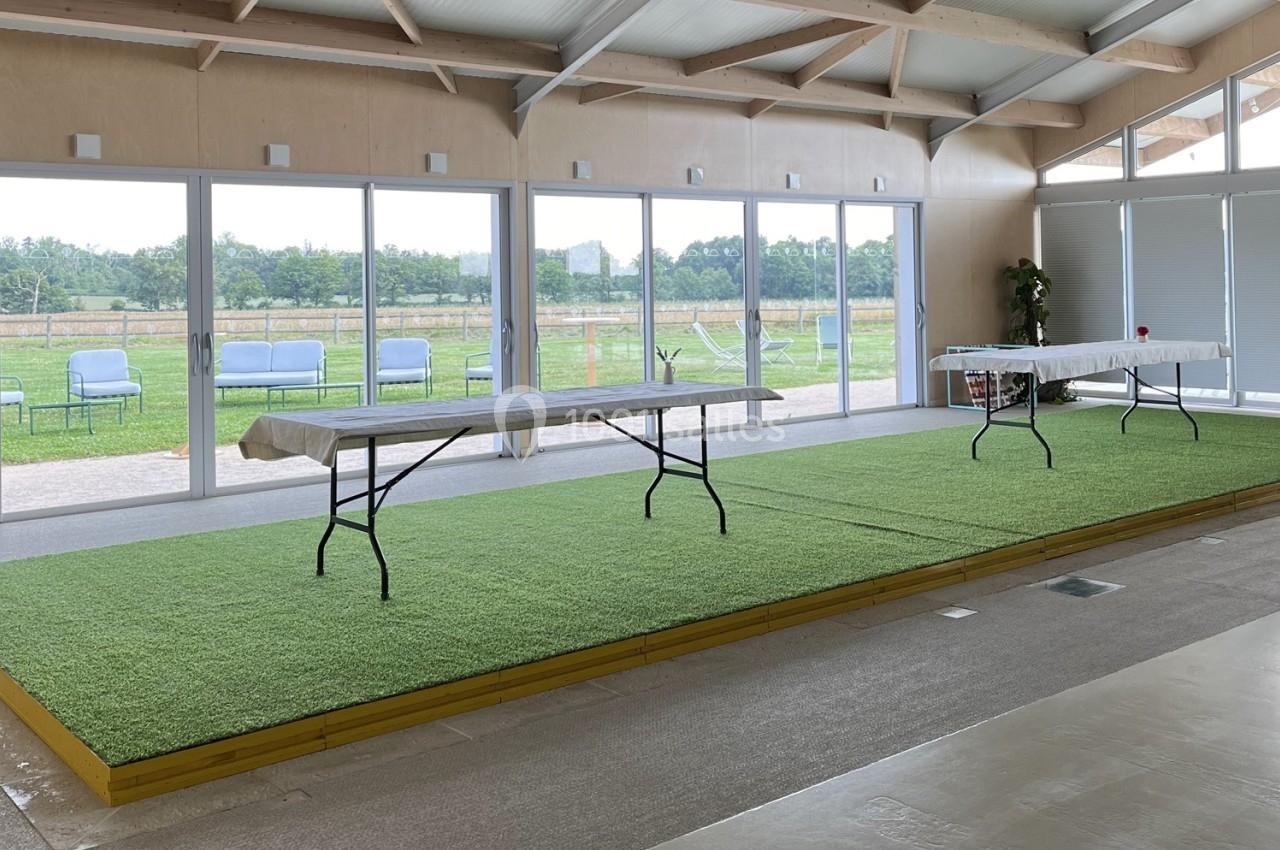 Salle lumineuse avec grandes baies vitrées, tables pliantes sur une estrade recouverte de gazon synthétique.