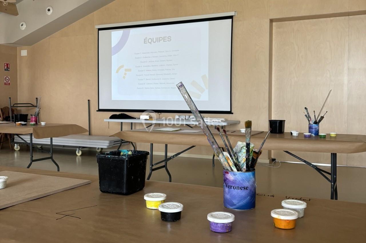 Salle d'atelier avec des tables recouvertes de papier kraft, des pinceaux, des pots de peinture et un écran affichant…