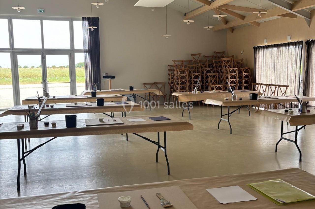 Salle lumineuse avec tables alignées, couvertes de matériel d'art, et des chaises empilées dans un coin.