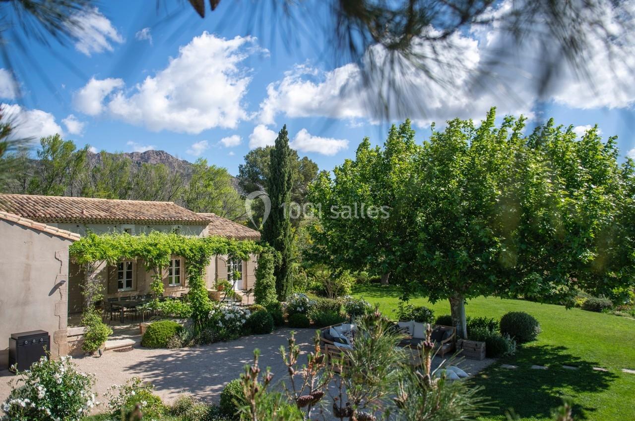 Maison en pierre entourée d'un jardin verdoyant avec arbres, arbustes et terrasse sous un ciel bleu.