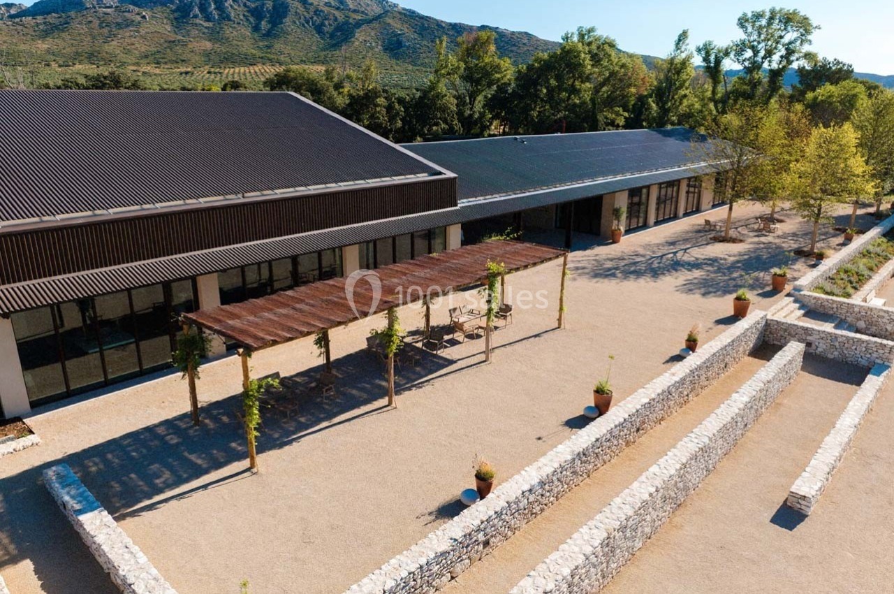 Vue aérienne d'un bâtiment moderne entouré de murs en pierre, avec une pergola et des montagnes en arrière-plan.