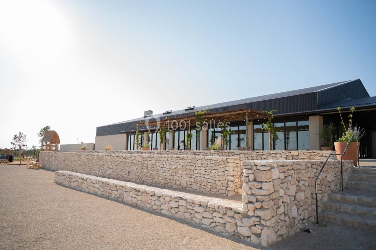 Bâtiment moderne avec terrasse ombragée, entouré de murs en pierre et situé dans un espace extérieur dégagé.