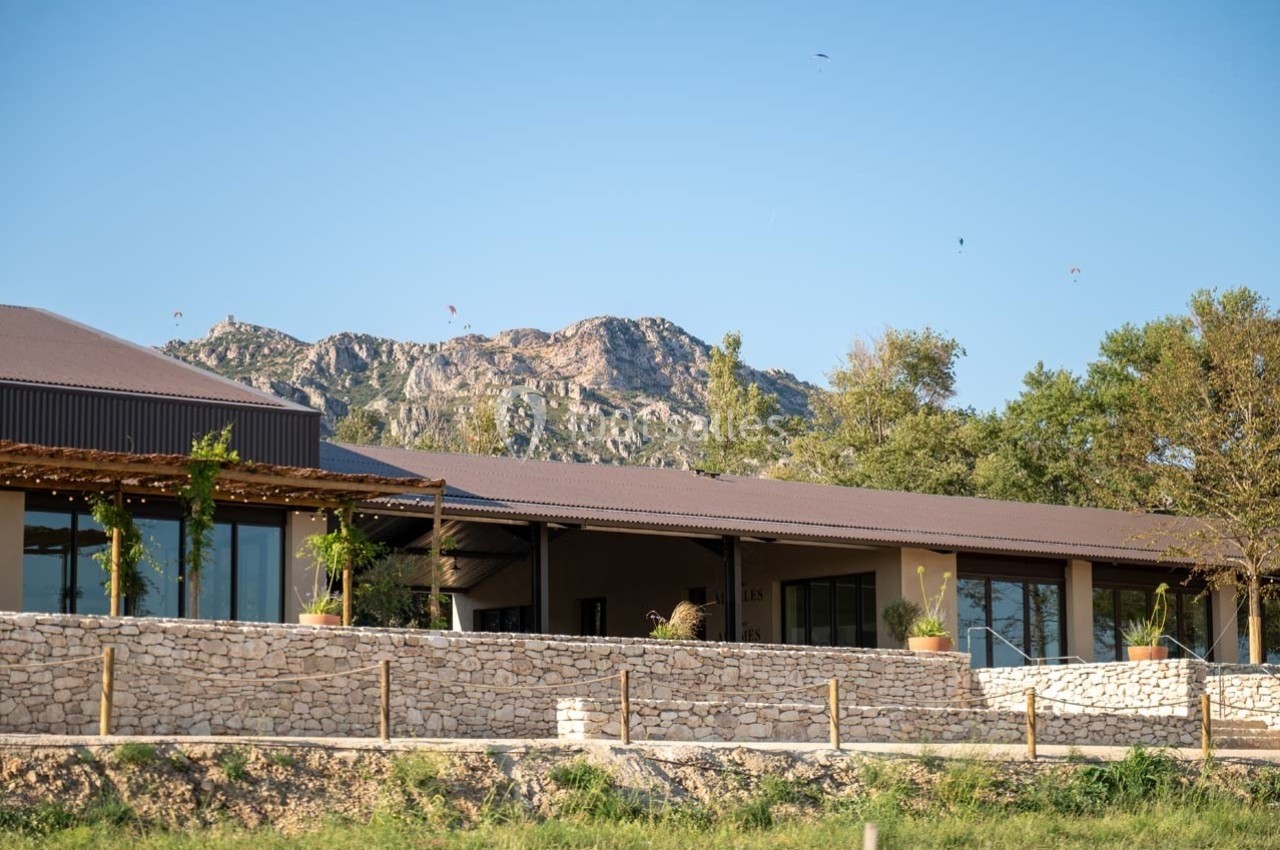 Bâtiment moderne avec terrasse en bois, entouré de végétation, au pied d'une montagne sous un ciel dégagé.