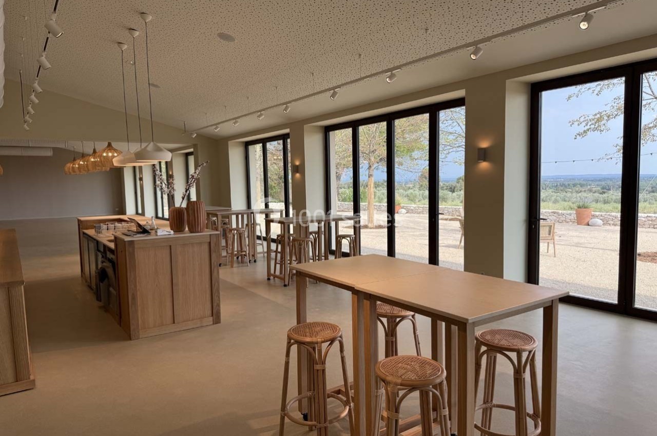 Salle lumineuse avec tables en bois, tabourets en rotin et grandes baies vitrées donnant sur un paysage extérieur.