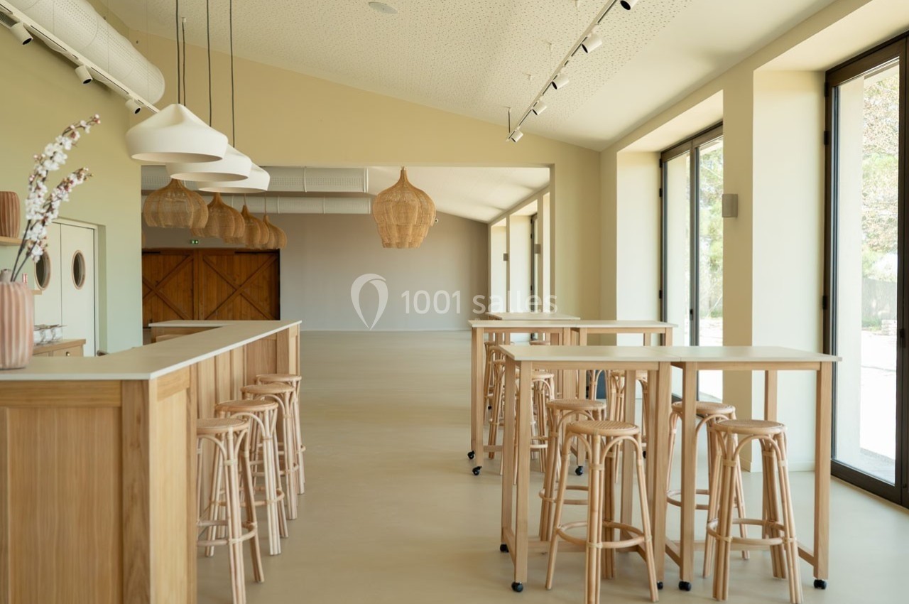 Salle lumineuse avec tables hautes en bois, tabourets assortis et suspensions en osier, grandes baies vitrées sur la droite.