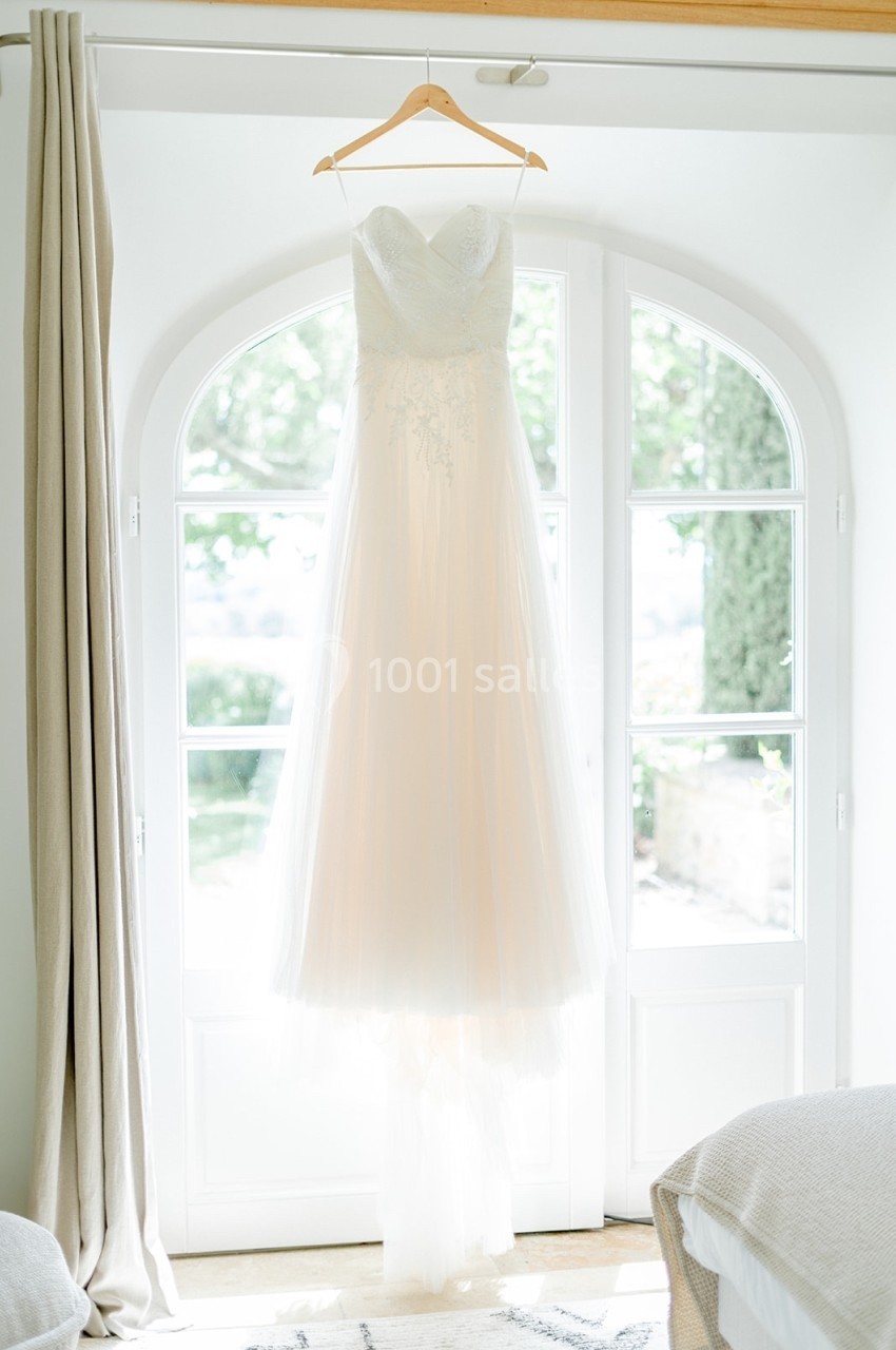 Robe de mariée blanche suspendue à un cintre devant une porte-fenêtre lumineuse avec vue sur un jardin.