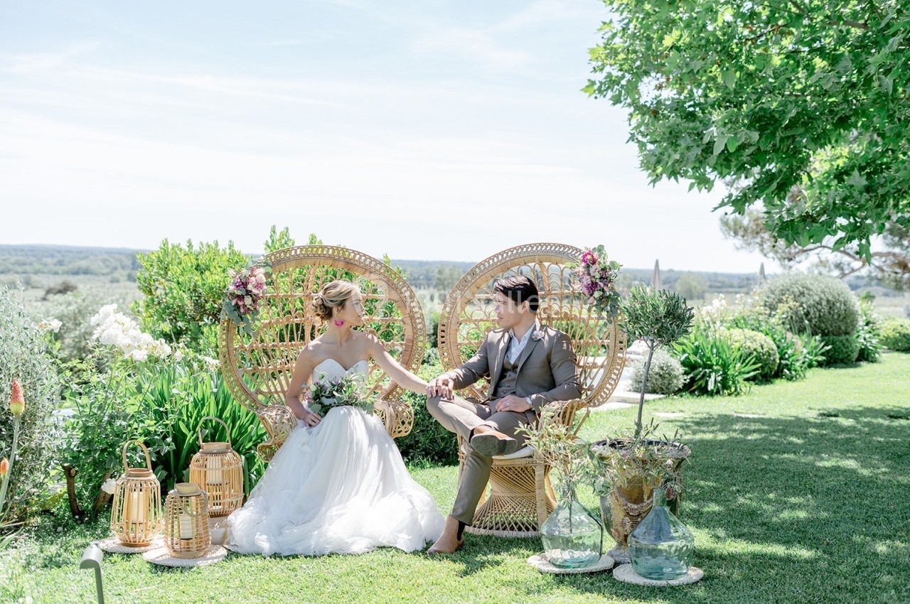 Un couple en tenue de mariage est assis sur des fauteuils en osier dans un jardin verdoyant et ensoleillé.