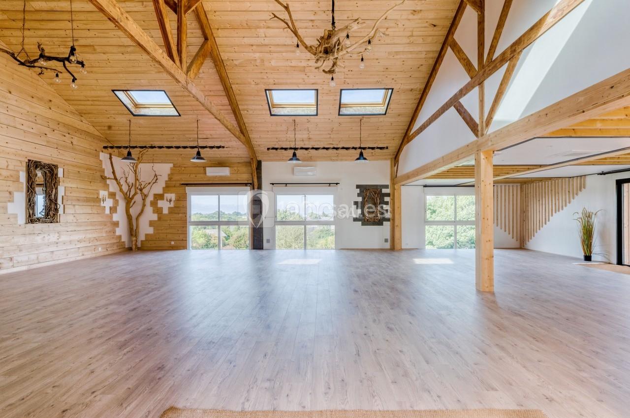 Salle spacieuse avec parquet clair, poutres apparentes en bois et grandes baies vitrées donnant sur un paysage verdoyant.