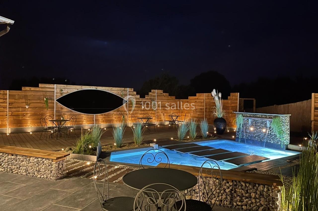 Terrasse en bois éclairée de nuit avec piscine, fontaine murale et mobilier de jardin entourés de plantes.