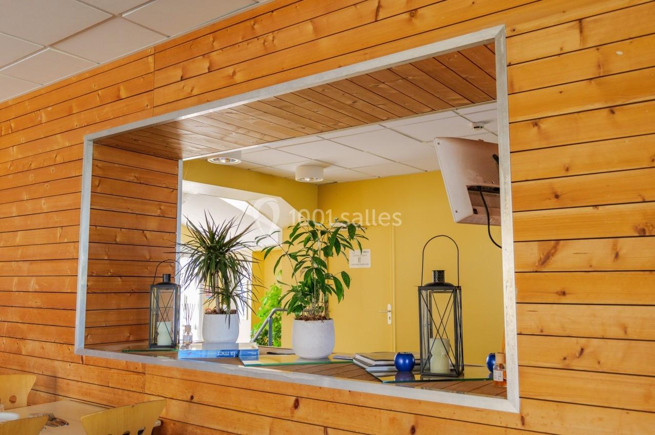 Vue à travers une ouverture dans un mur en bois sur un espace intérieur avec plantes, lanternes et mur jaune.