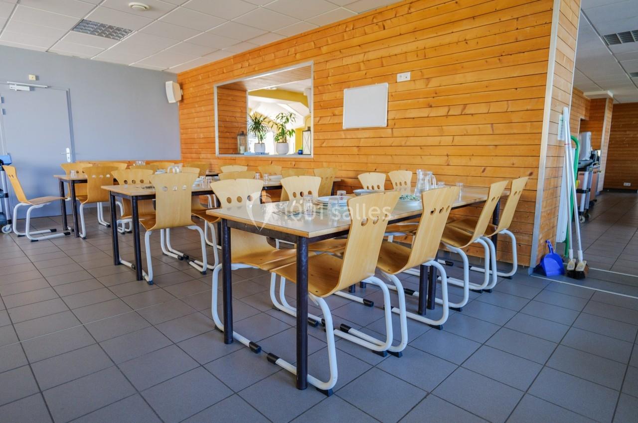 Salle de restauration avec tables en bois clair, chaises assorties et vaisselle disposée, murs en bois et carrelage gris.