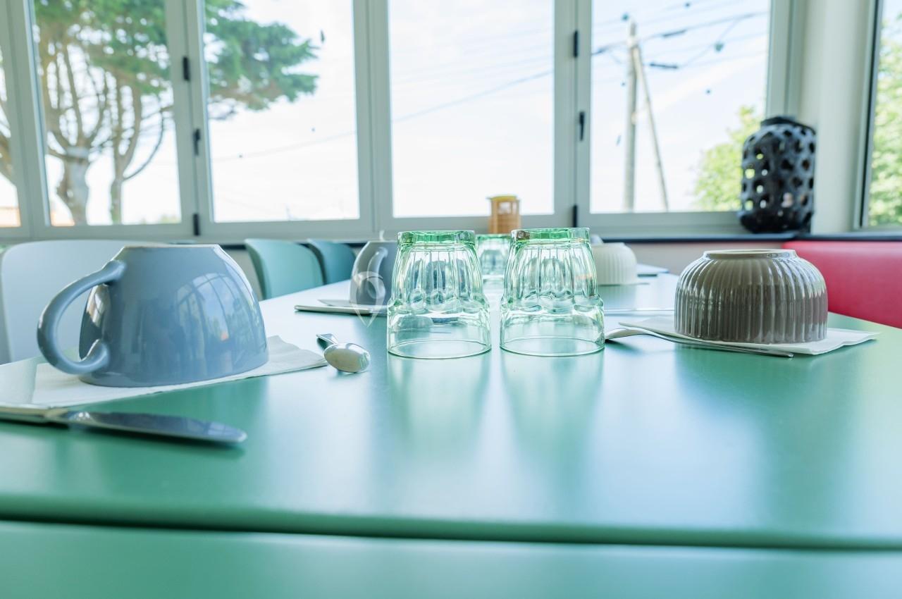 Table dressée avec des tasses, verres retournés, couverts et serviettes, près d'une baie vitrée donnant sur l'extérieur.