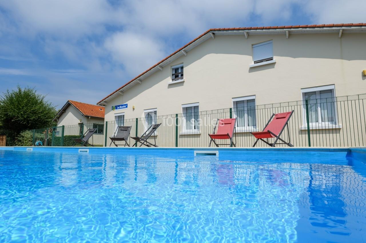 Piscine extérieure avec eau bleue claire, entourée de chaises longues colorées devant une maison beige.