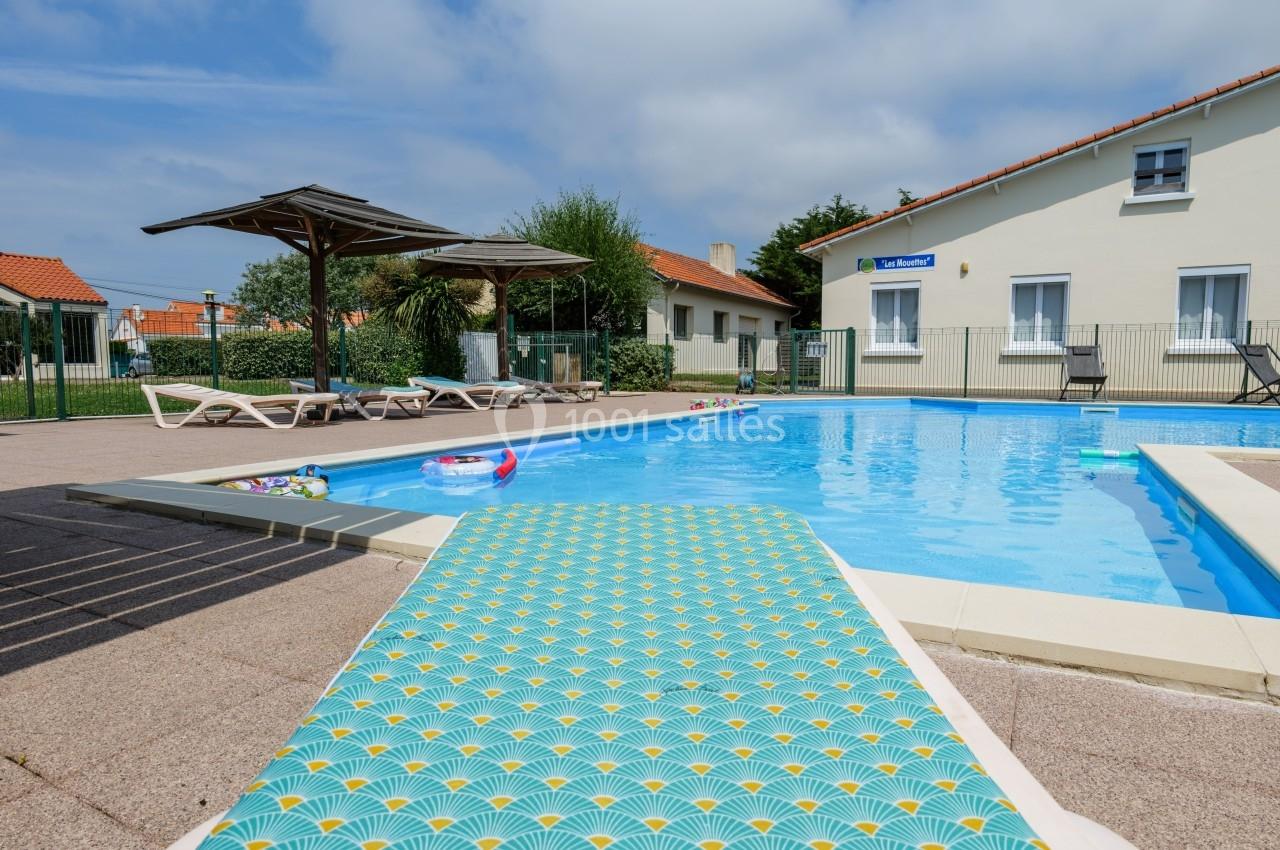 Piscine extérieure avec transats, parasols et jouets flottants, entourée de maisons sous un ciel partiellement nuageux.