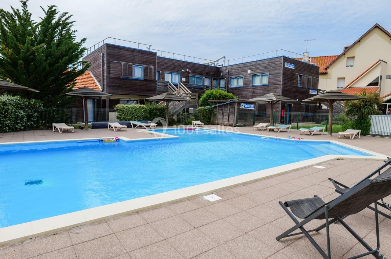 Piscine extérieure entourée de chaises longues, située devant des bâtiments en bois et des maisons avec toits en tuiles.