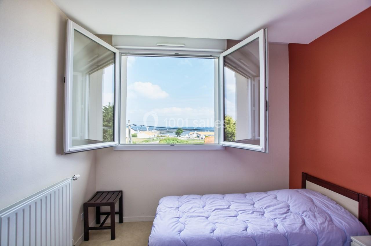 Fenêtre ouverte dans une chambre avec lit simple, vue sur la mer et maisons au loin.