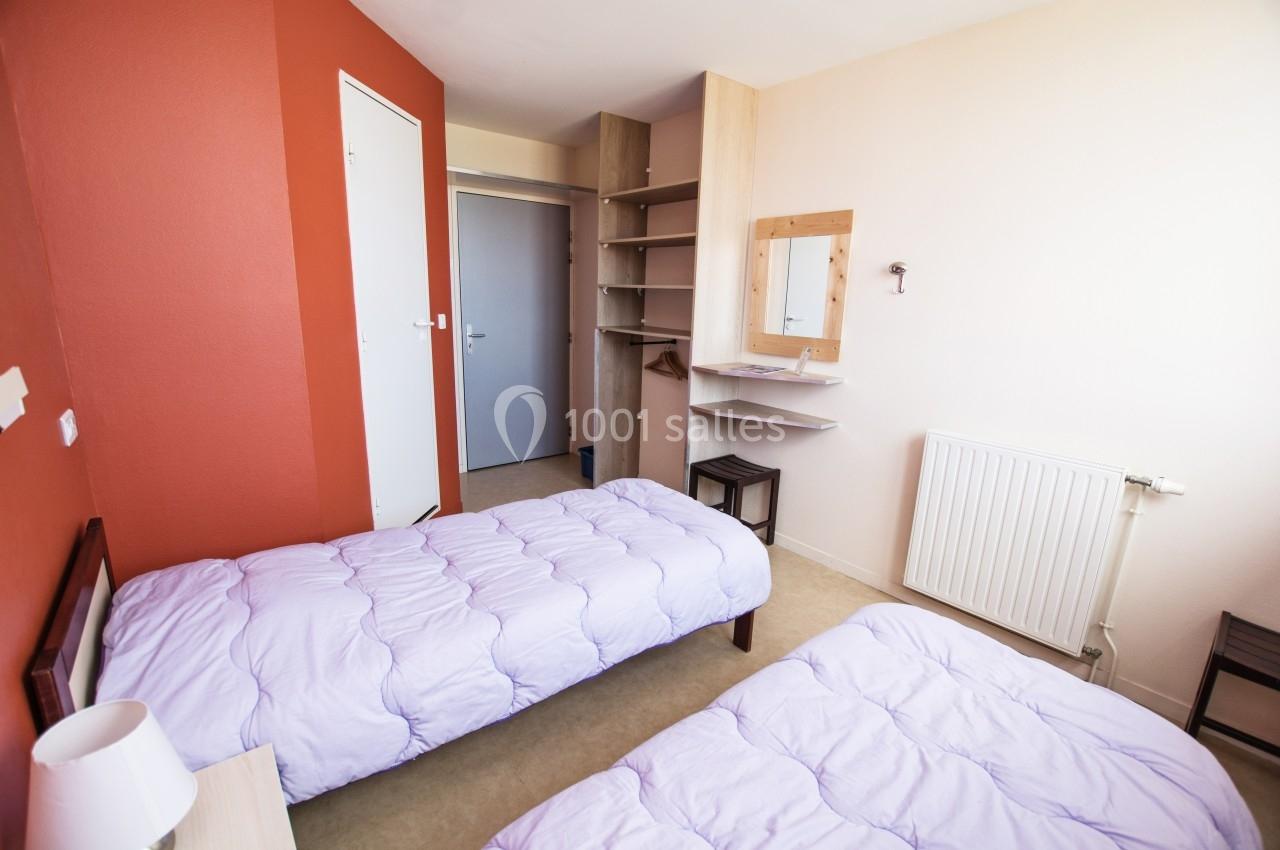 Chambre simple avec deux lits simples, murs rouges et blancs, étagères, miroir, et porte ouverte sur un couloir.